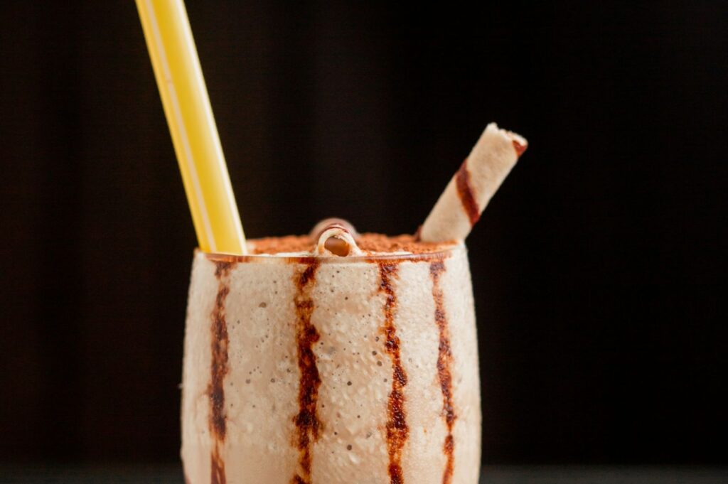 Creamy milkshake in a glass with chocolate drizzle, topped with a striped wafer stick and a yellow straw. Rich, indulgent, and inviting.