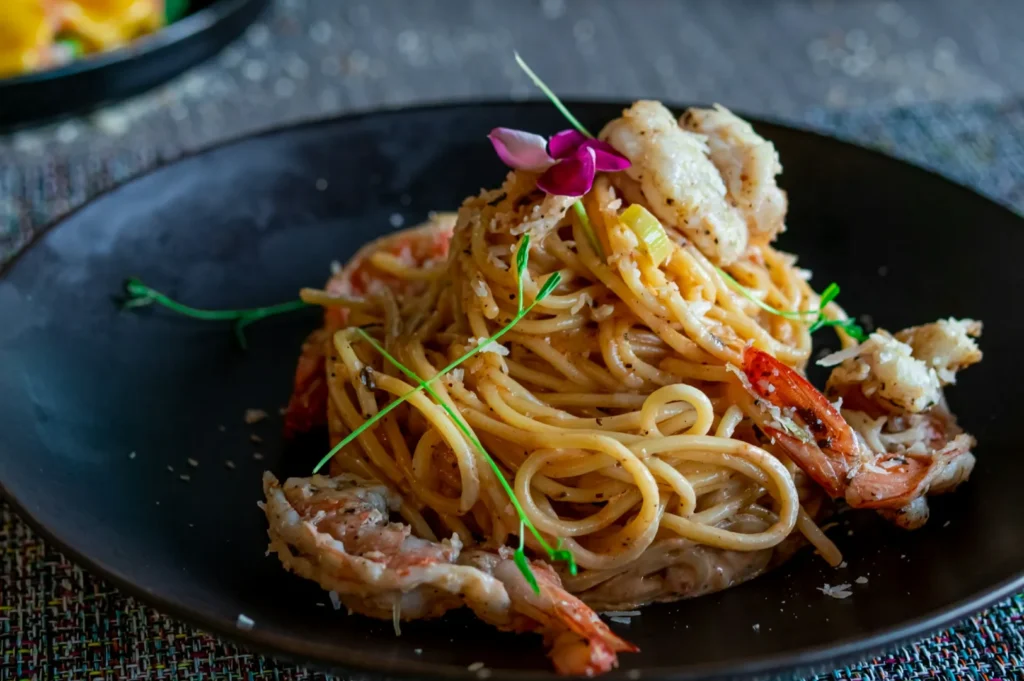 A black plate with creamy spaghetti topped with shrimp, a sprinkle of herbs, and a pink flower garnish, evoking a sophisticated dining experience.