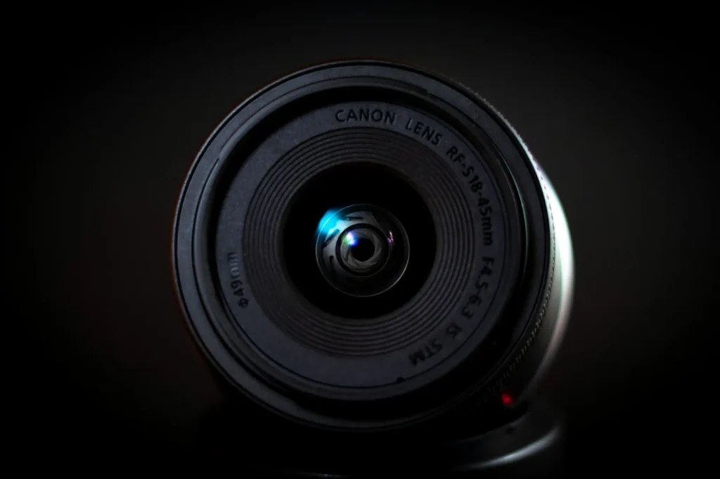 Close-up of a Canon camera lens facing forward, illuminated by a soft blue light, set against a dark background. The focus is sharp and centered.