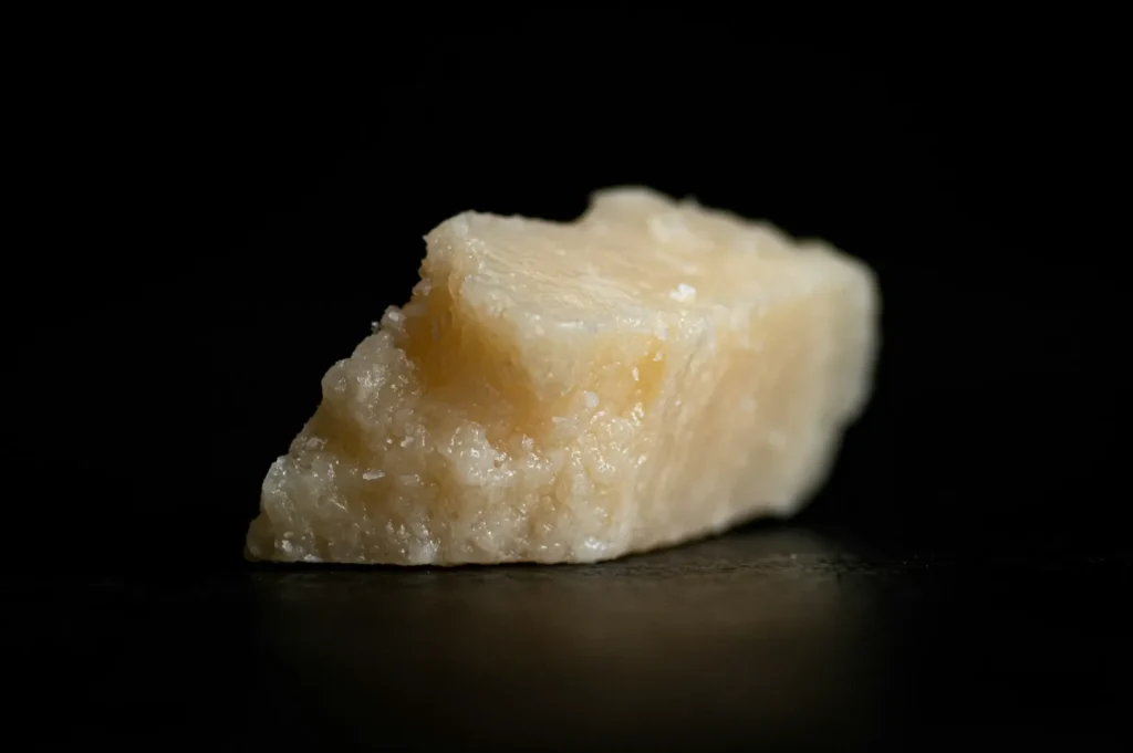 A close-up of a piece of Parmesan cheese on a black background, showing its rough, crumbly texture and pale yellow color with subtle lighting