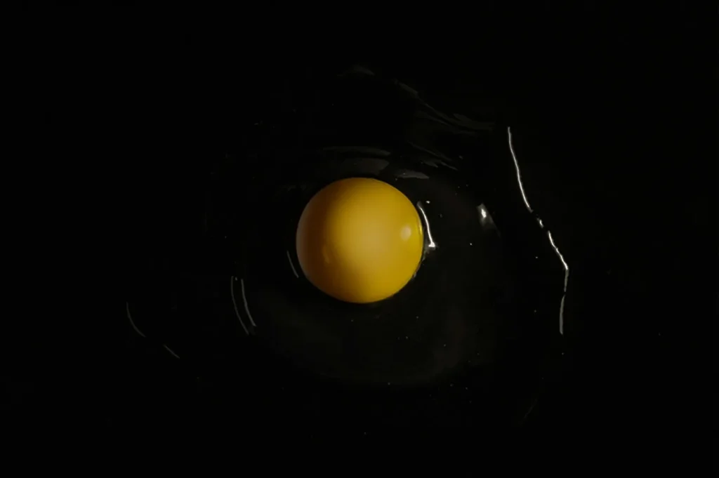 A single egg yolk surrounded by clear egg white on a black background. The lighting highlights the yolk's smooth texture, creating a minimalist, elegant look.