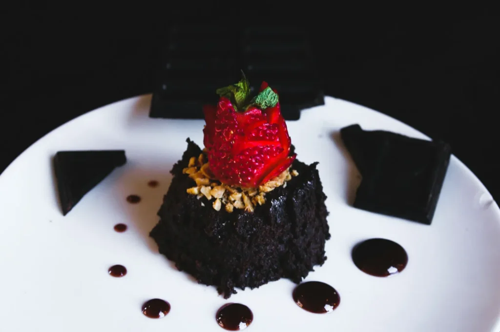 Decadent chocolate cake with a textured top, garnished with a vivid red strawberry and nuts, drizzled with syrup on a white plate.