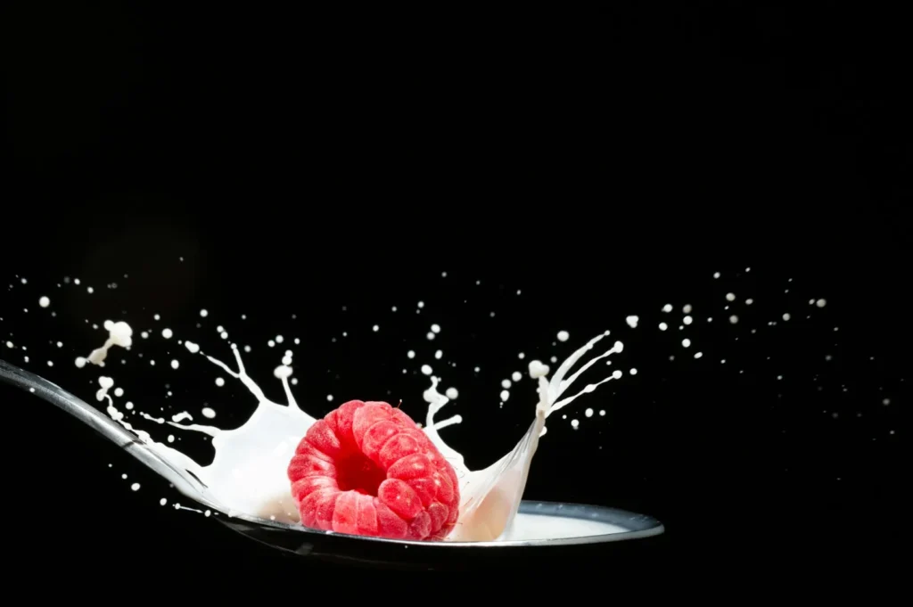 “A vibrant red raspberry splashes into milk, causing droplets to fly in all directions against a dark background, creating a dynamic and fresh scene.”