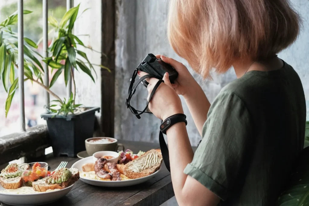 A person with short hair and a green shirt photographs a table with plates of brunch food, including toast and a drink, near a window with a potted plant.
