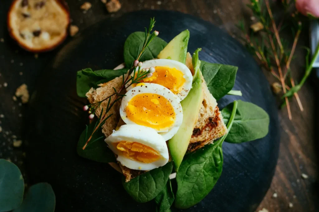 Open-faced sandwich with spinach, sliced egg, and avocado on toasted bread. Garnished with herbs, served on a dark plate, rustic and fresh.