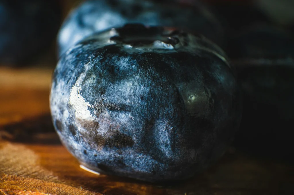 Close-up of a plump blueberry with a deep blue, glossy surface and water droplets. It sits on a wooden surface, conveying freshness and juiciness.