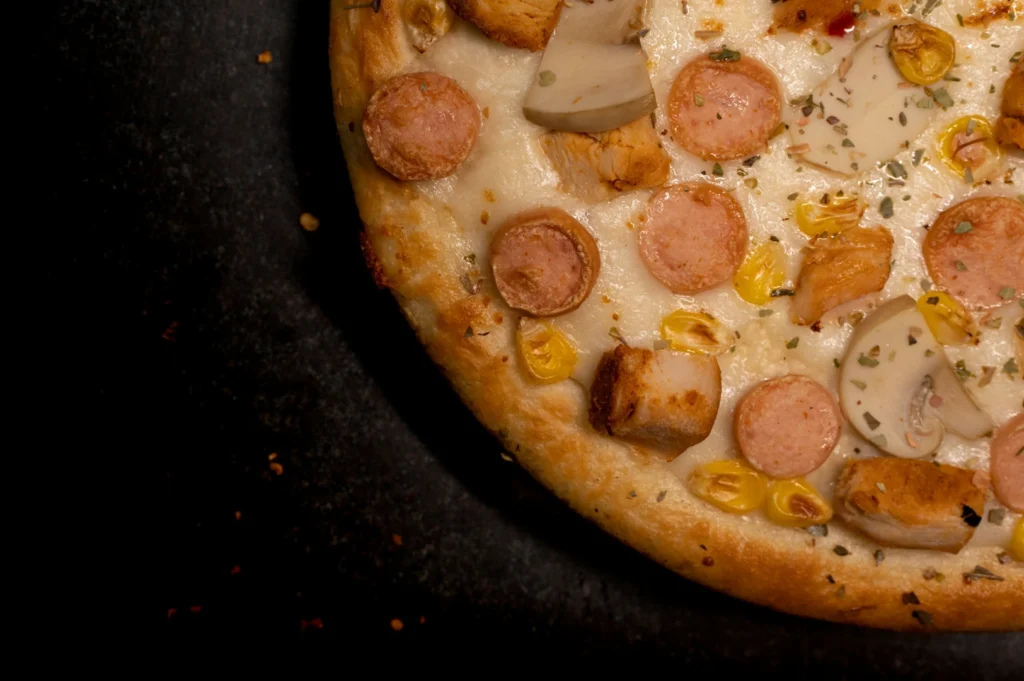 Close-up of a pizza with a golden crust topped with sausage slices, mushrooms, corn, chicken pieces, and herbs, set against a dark background.