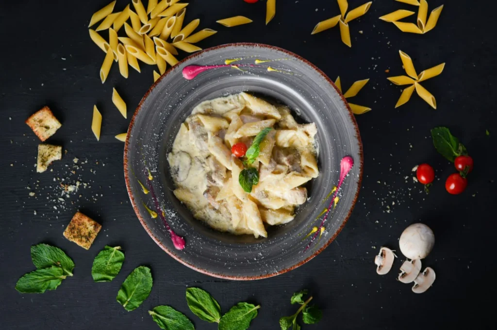 A bowl of creamy penne pasta with mushrooms and herbs sits on a dark surface, surrounded by uncooked pasta, cherry tomatoes, mushrooms, and fresh mint leaves, creating a gourmet and artistic presentation.