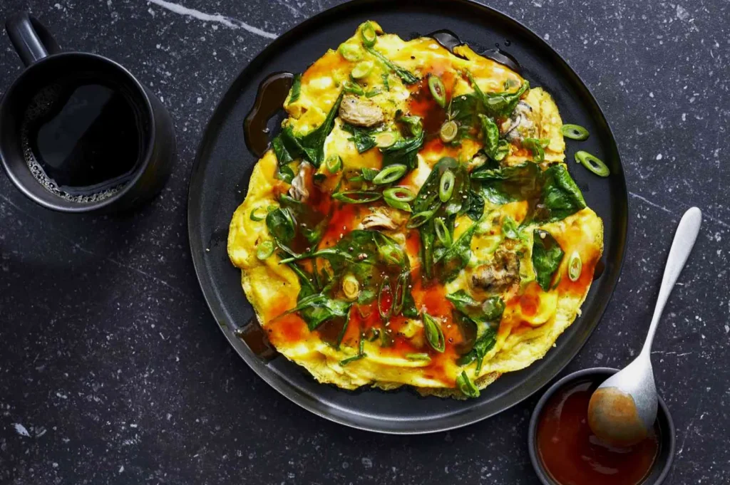 A vibrant omelet with spinach and green onions, topped with red sauce on a black plate. Beside it, a cup of coffee and a small dish with a spoon.