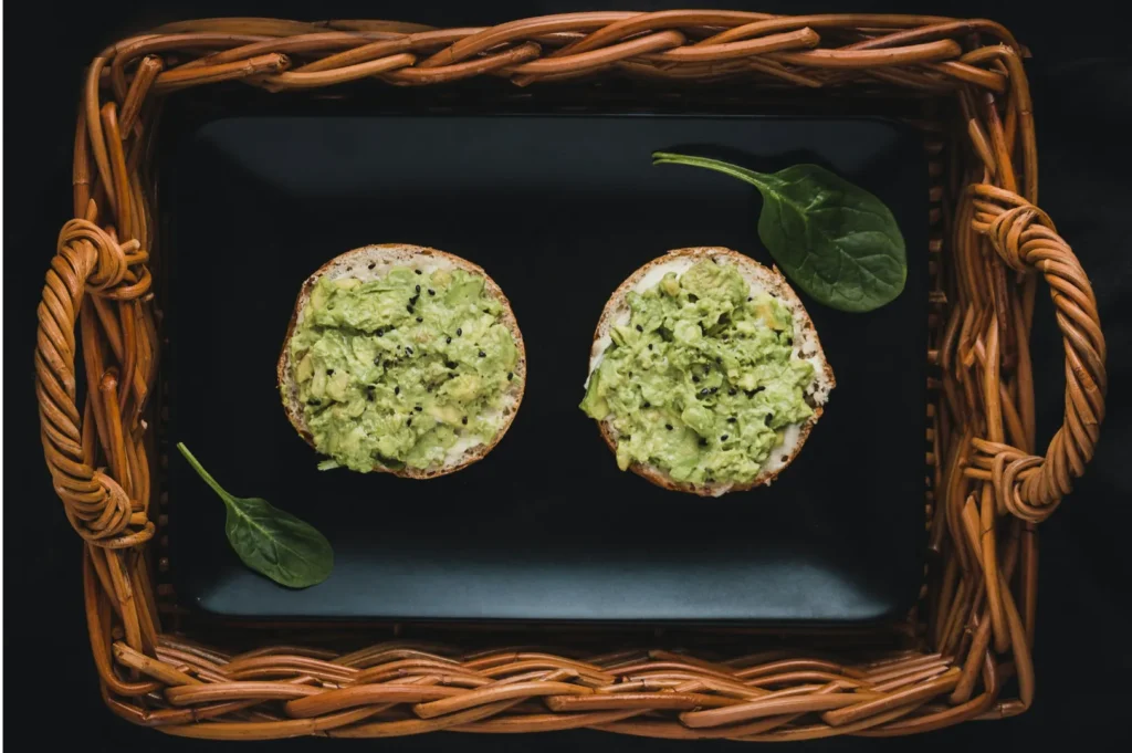 Two bagel halves with avocado spread on a black plate, placed in a wicker basket. Spinach leaves rest beside them, creating a fresh, rustic vibe.