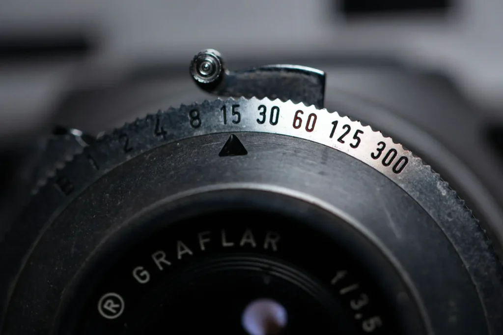 Close-up of a vintage camera lens, focusing on the aperture dial with numbers ranging from B to 300 and the word “GRAFAR” visible on the lens.