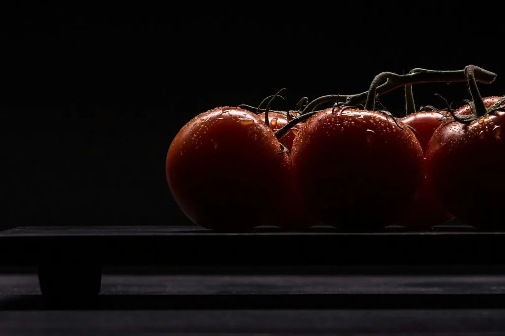 “A cluster of fresh red tomatoes on a vine sits on a dark wooden surface. Water droplets glisten on the tomatoes, set against a black background.”