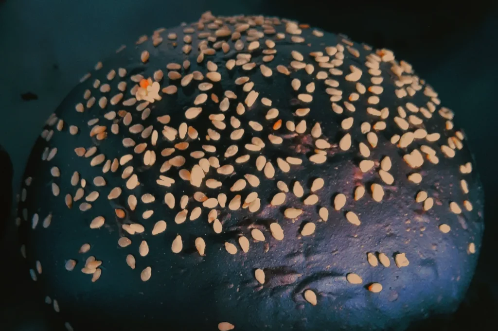 “Close-up of a black sesame seed bun with an even distribution of seeds on top. The bun has a glossy texture set against a dark background.”