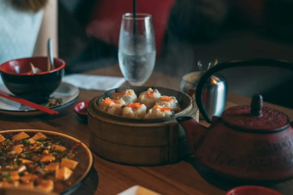 Steaming dim sum in a bamboo basket is surrounded by a red teapot, a glass of water, and dishes with chopsticks on a wooden table. Cozy dining atmosphere.