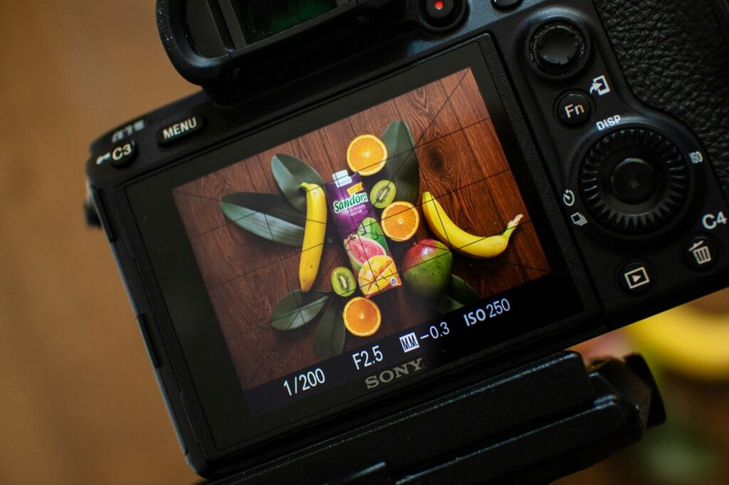 Close view of the camera display capturing a carefully arranged fruit scene, featuring a Sandora beverage carton, ripe bananas, halved oranges, kiwi, and glossy green leaves on a wooden surface.