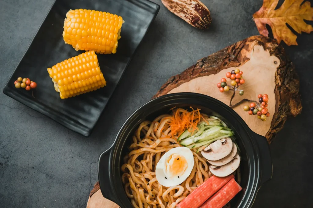 A bowl of seasoned noodles with a halved boiled egg, crab sticks, mushrooms, carrots, and cucumber, alongside corn on a black plate, on a rustic wood board.