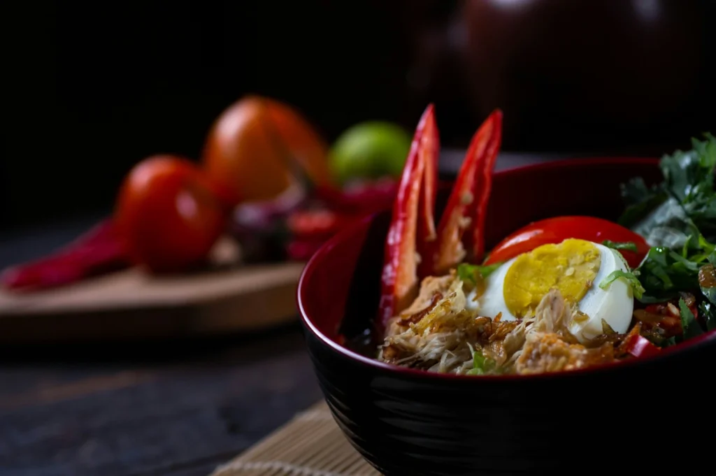 A vibrant bowl of noodles with sliced boiled egg, red chili, and greens, set against a dark background. Fresh tomatoes and limes blur in the backdrop.
