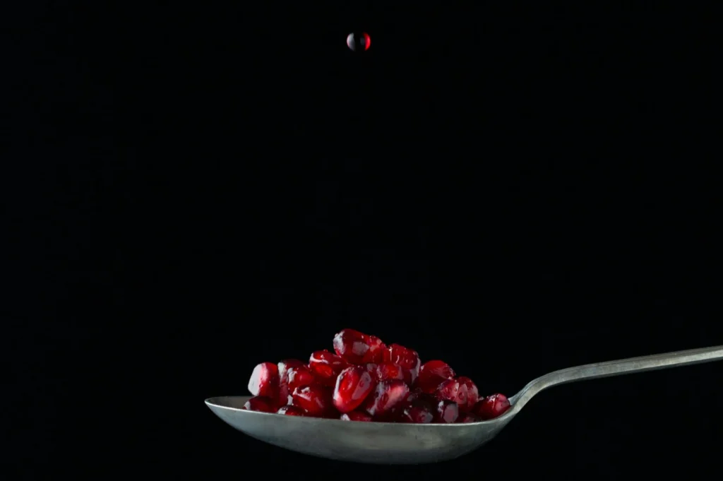 A silver spoon holds vibrant red pomegranate seeds against a black background, with a single seed hovering above, creating a sense of motion and focus.