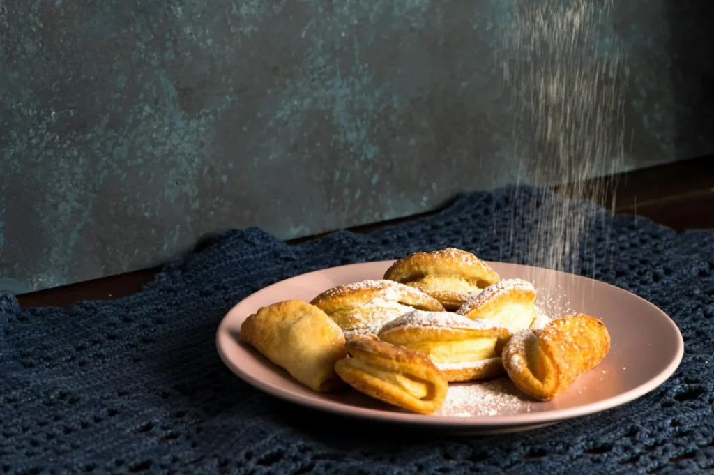 A pink plate with golden-brown pastries, dusted with powdered sugar, on a dark blue textured cloth. Sugar falls, adding a touch of elegance.