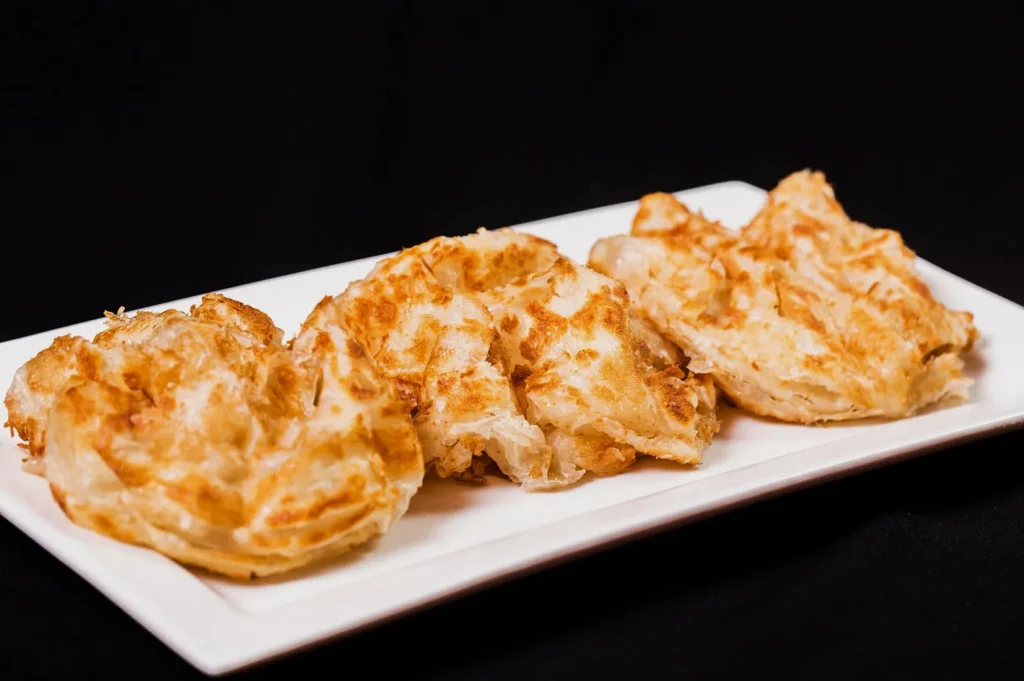 Three crispy, golden-brown parathas on a rectangular white plate, set against a dark background. The parathas appear flaky and freshly cooked.