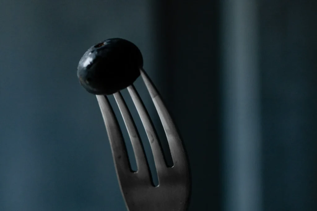 “A single blueberry is pierced on a silver fork against a dark blue, moody background. The scene conveys simplicity and focus.”