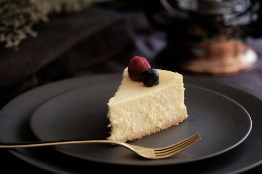 A pale cheesecake slice with a crumbly crust sits on a dark plate, topped with a raspberry and a blueberry, with a gold fork nearby.