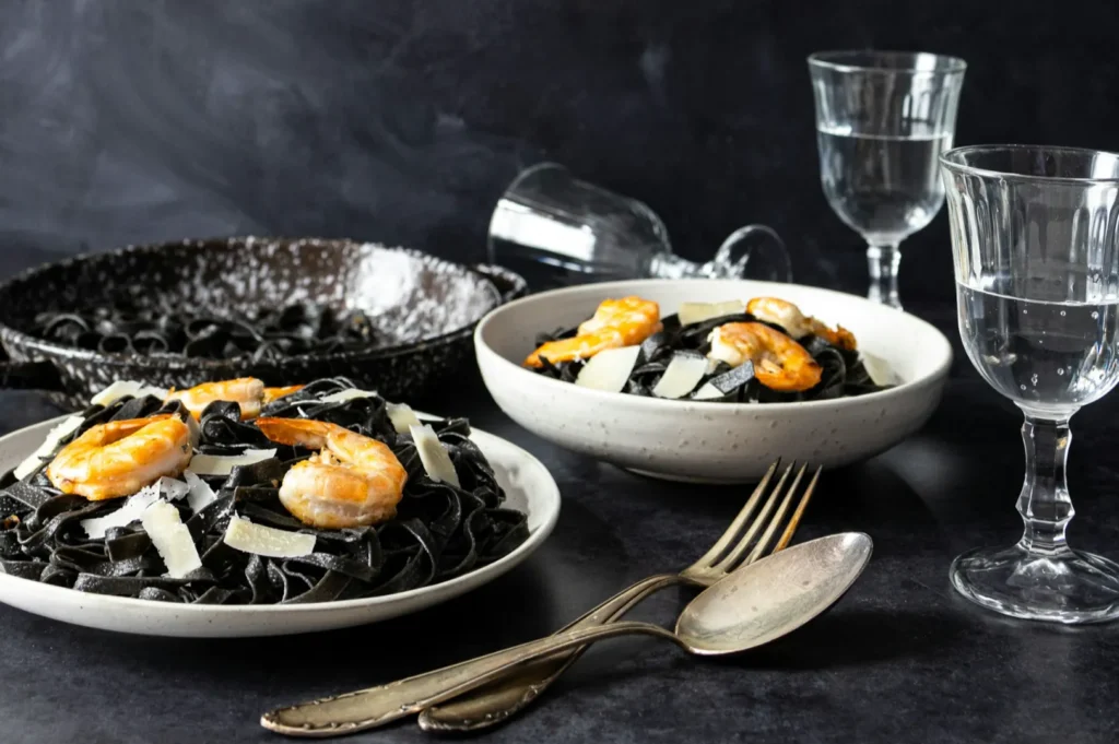Elegant table setting with two plates of black pasta topped with shrimp and Parmesan, surrounded by glassware and utensils on a dark surface.