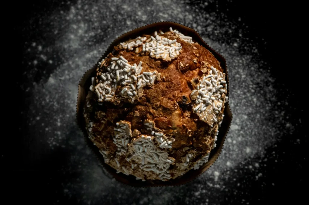 Top-down view of a panettone, a round, golden-brown Italian cake with white sugar crystals, on a dark background dusted with flour.