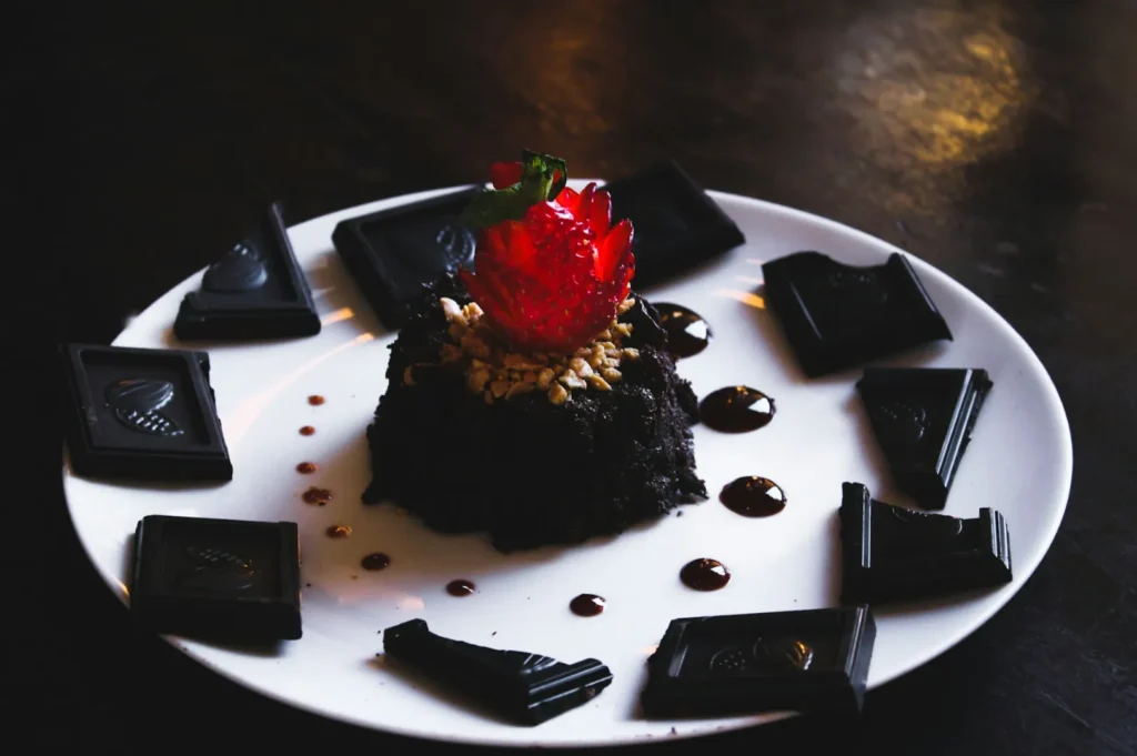 A dessert featuring a dark chocolate cake garnished with a sliced strawberry and nuts, surrounded by chocolate pieces on a white plate.