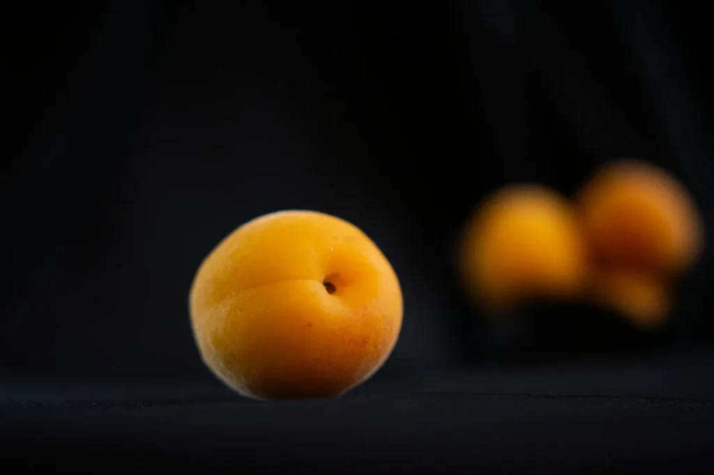 “A single apricot is sharply focused against a black background, while two blurry apricots are in the distance, creating a dramatic contrast.”