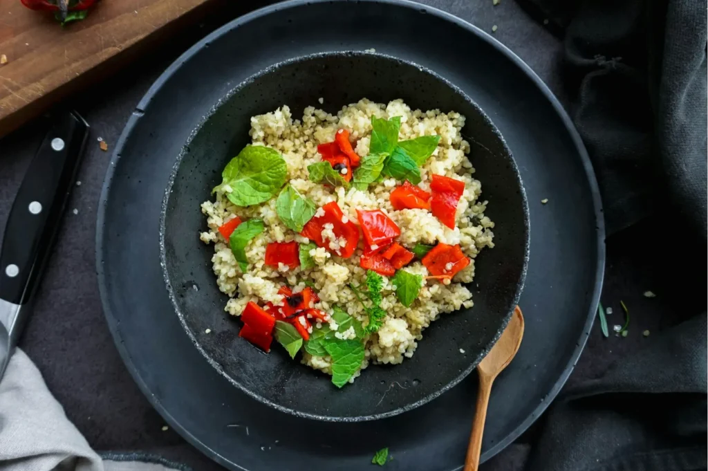 A black plate with a quinoa salad topped with bright red bell pepper pieces and fresh green mint leaves, set on a dark table with a wooden spoon.