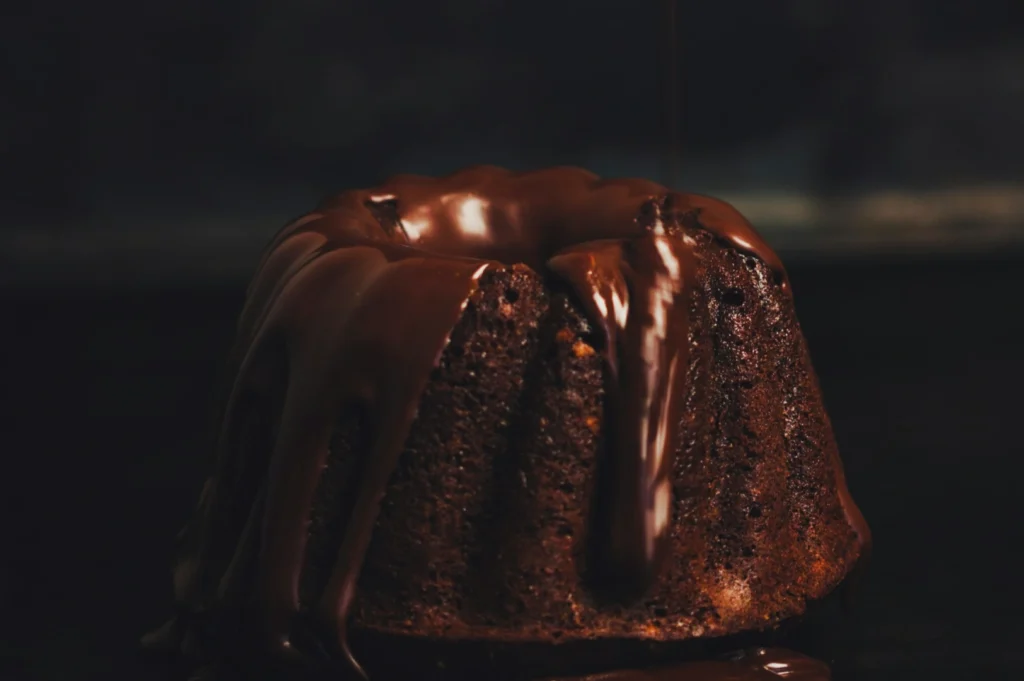 A rich chocolate bundt cake with glossy melted chocolate drizzled over the top, creating a decadent and indulgent appearance. Dark, moody background.