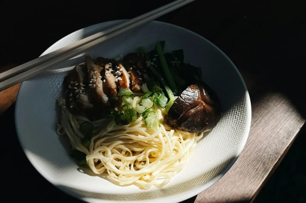 A white bowl in warm lighting holds noodles, sliced chicken with sesame seeds, green vegetables, and a shiitake mushroom. Chopsticks rest on the rim.