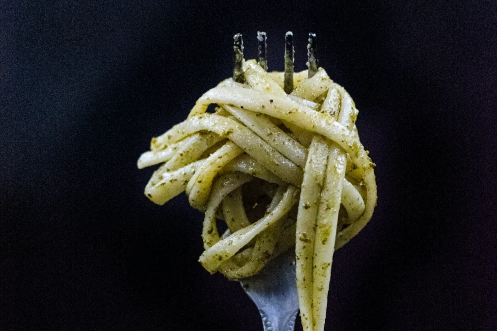 Close-up of a fork holding tangled linguine coated in green pesto sauce against a dark background, conveying a sense of savory deliciousness.