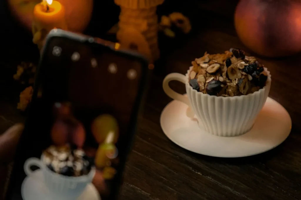 A white teacup filled with oat-topped dessert sits on a saucer beside a candle, creating a warm, cozy ambiance. A smartphone captures the scene.