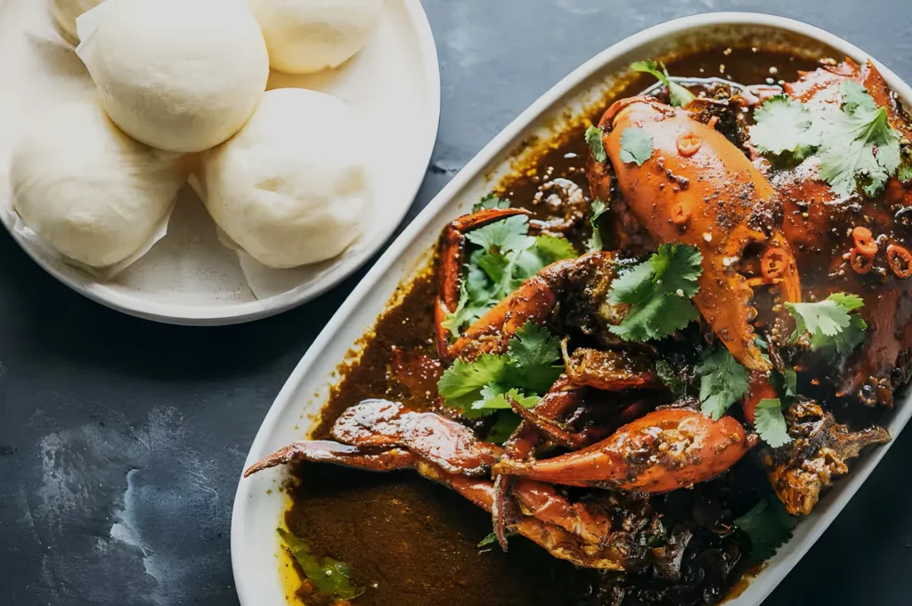 A white plate holds spicy crab curry garnished with cilantro, next to a bowl of steamed buns on a dark surface, creating a bold, flavorful presentation.
