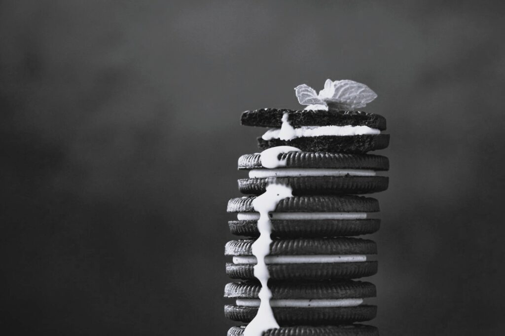 A tall stack of chocolate sandwich cookies with generous cream filling leaking between layers, finished with a delicate mint leaf on top, in monochrome.