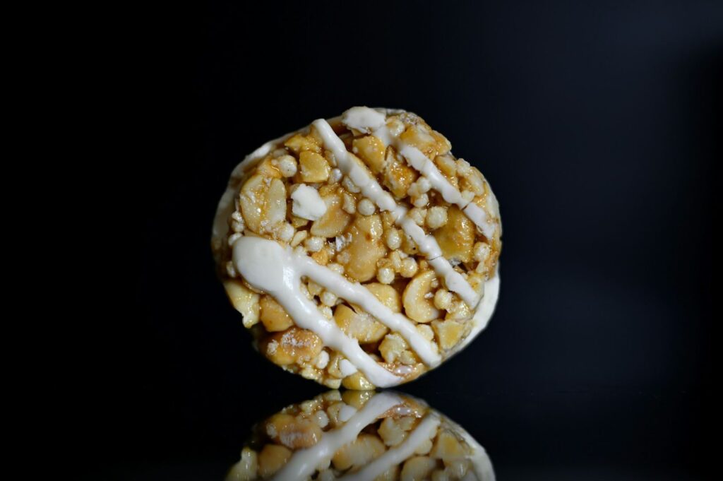 Round peanut brittle with white icing drizzles on a glossy black surface, reflecting underneath. The scene conveys a sense of sweetness and indulgence.