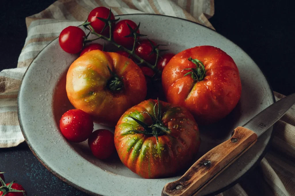 A rustic plate holds three heirloom tomatoes sprinkled with water droplets, surrounded by cherry tomatoes. A knife lies beside, on a striped cloth.