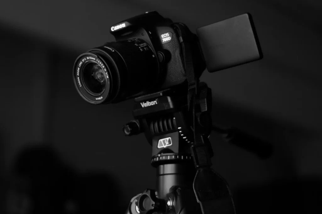 A black and white photo of a DSLR camera mounted on a tripod. The camera, angled slightly to the left, has a lens cap attached. The mood is professional.