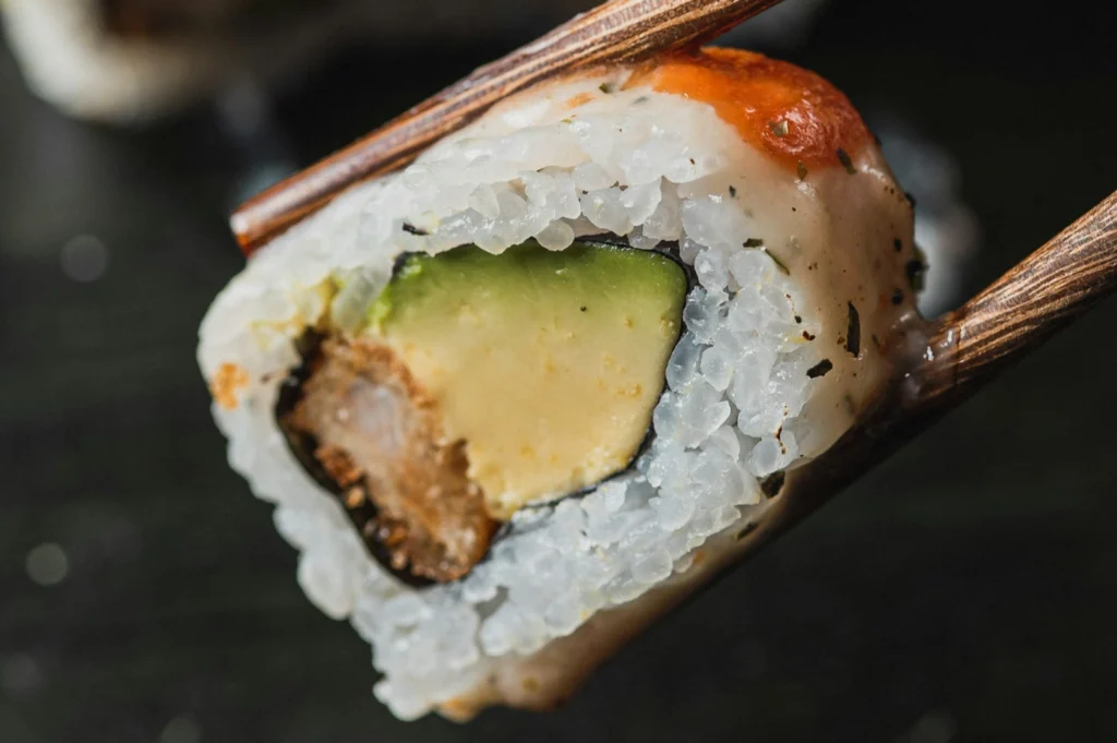 Close-up of a sushi roll with avocado, crab, and rice, held by chopsticks. The textured surface and vibrant colors convey freshness.