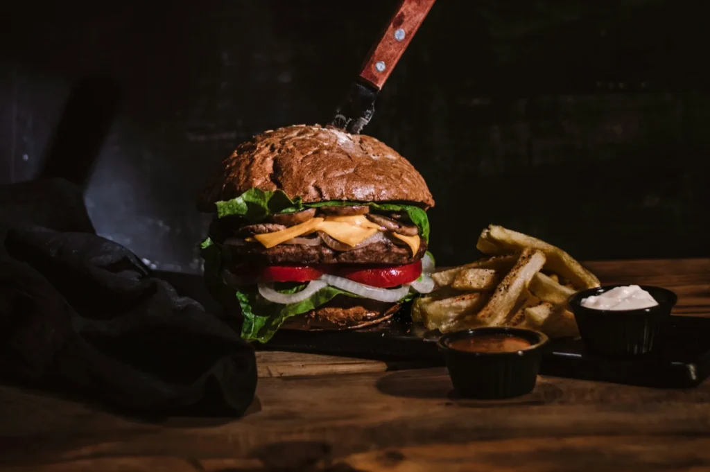 A rustic burger with lettuce, tomato, cheese, and onion is pierced by a knife, accompanied by fries and dipping sauces on a wooden table, creating a bold, hearty scene.