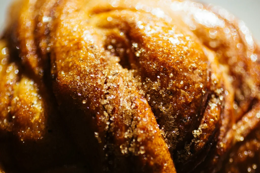 Close-up of a freshly baked pastry with golden-brown crust, glistening sugar crystals, and a textured surface, evoking warmth and sweetness.