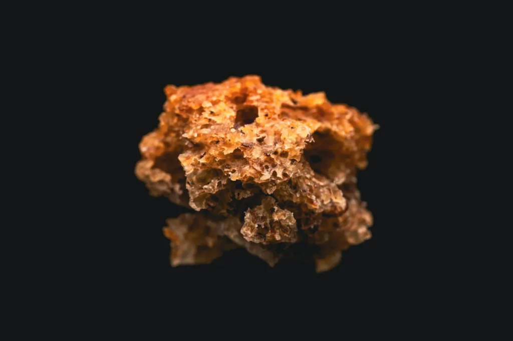 A caramel-colored honeycomb toffee piece sits on a shiny black surface, displaying its porous texture and casting a subtle reflection below.