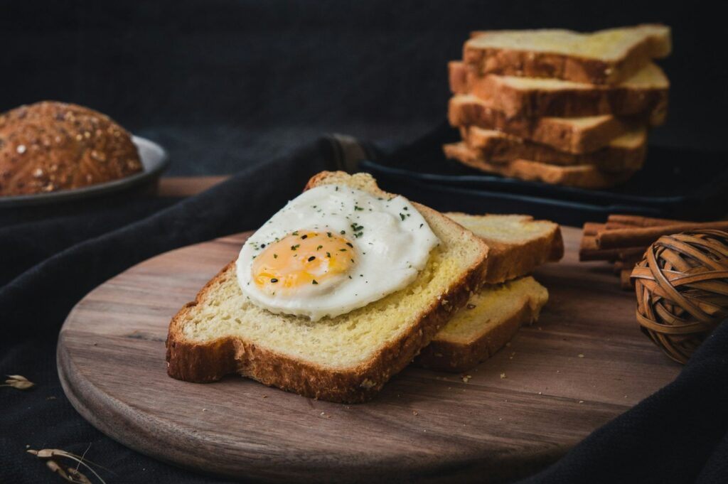 A close-up of a thick slice of golden toast topped with a sunny-side-up egg, sprinkled with herbs, placed on a wooden board with more bread in the softly lit background.
