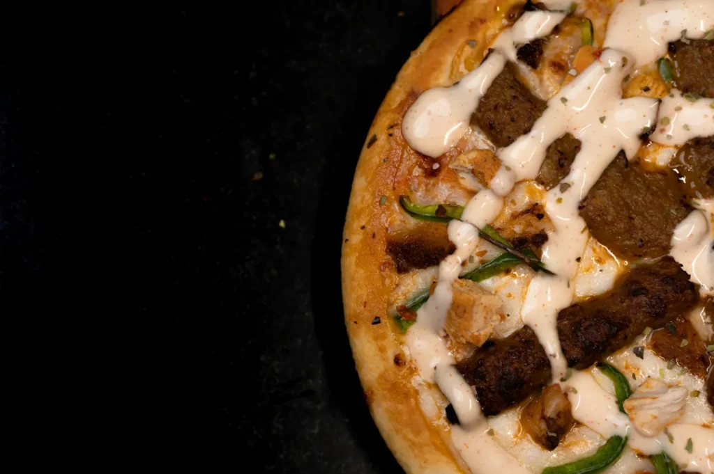 Close-up of a pizza featuring sausage slices, chicken pieces, green peppers, and white sauce drizzles, on a dark background.