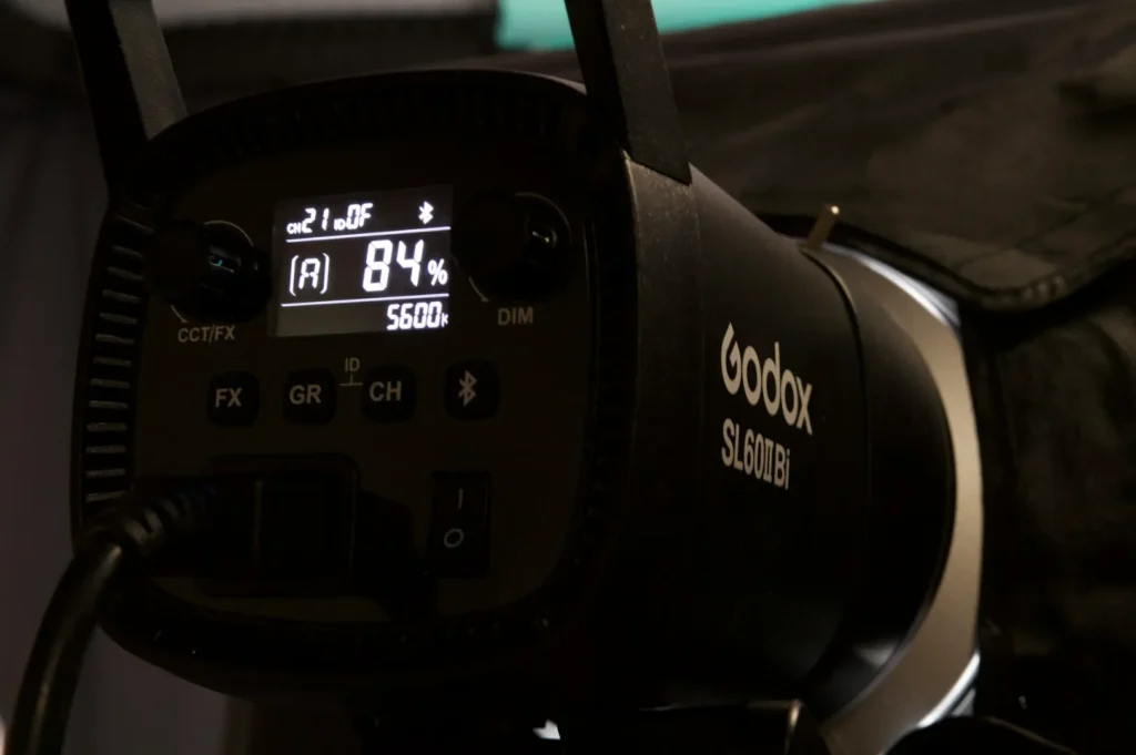 A close-up of a Godox SL60D video light displaying settings, including an 84% brightness at 5600K. The device is in a dimly lit studio setting.