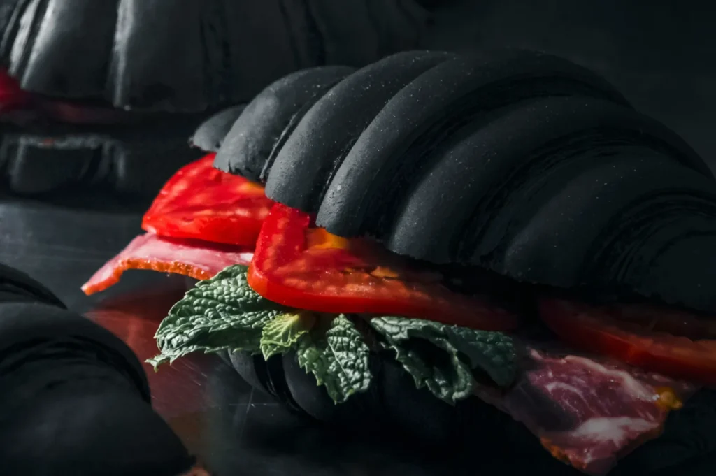 A black croissant sandwich with fresh tomatoes, lettuce, and cured meat is displayed on a dark surface, creating a bold and mysterious culinary vibe.