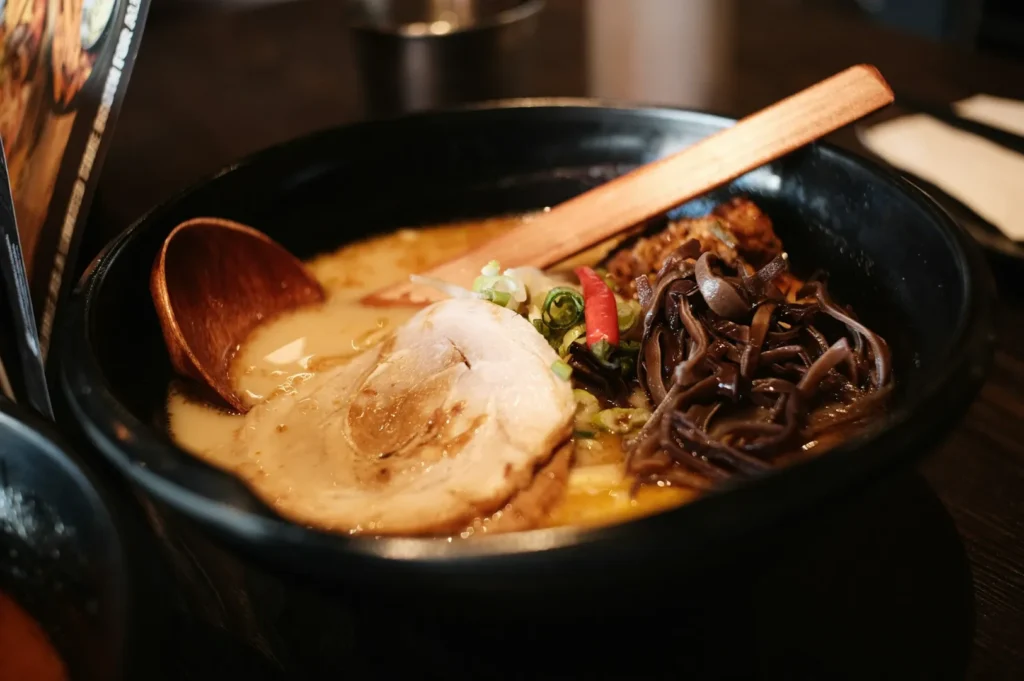 A black bowl of ramen with creamy broth, topped with a slice of pork, green onions, wood ear mushrooms, and a wooden spoon, creating a warm, savory tone.