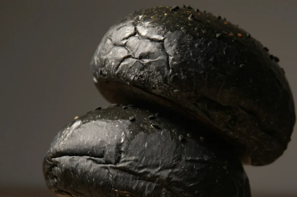 Close-up of two stacked black hamburger buns with a glossy finish and scattered sesame seeds. The surface is wrinkled, creating a textured appearance.